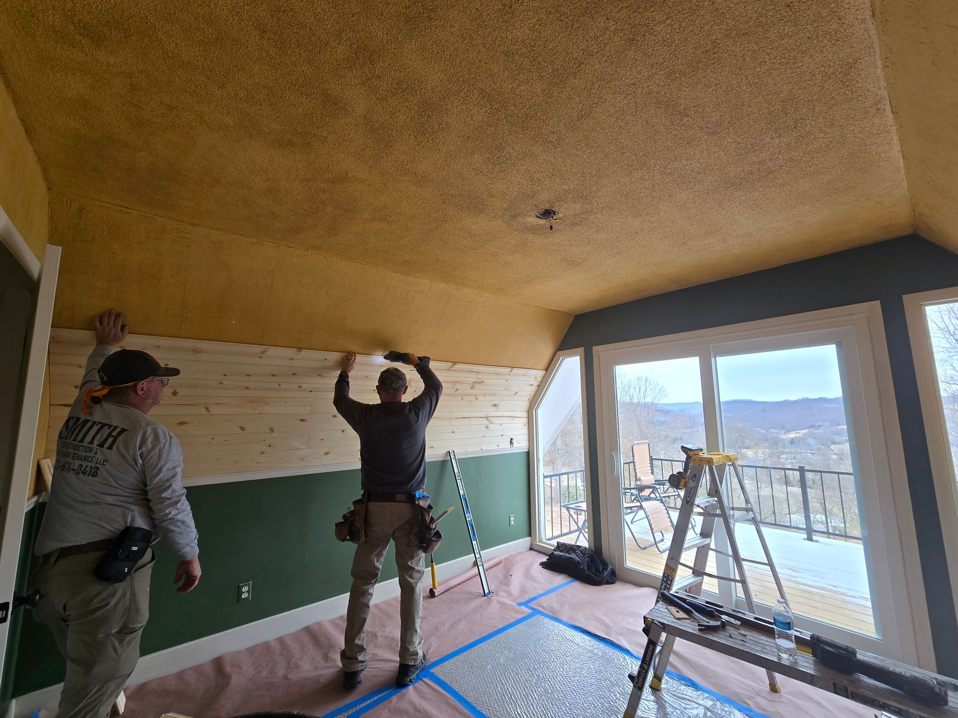 Two workers plastering a room with a curved ceiling and large windows overlooking mountains.