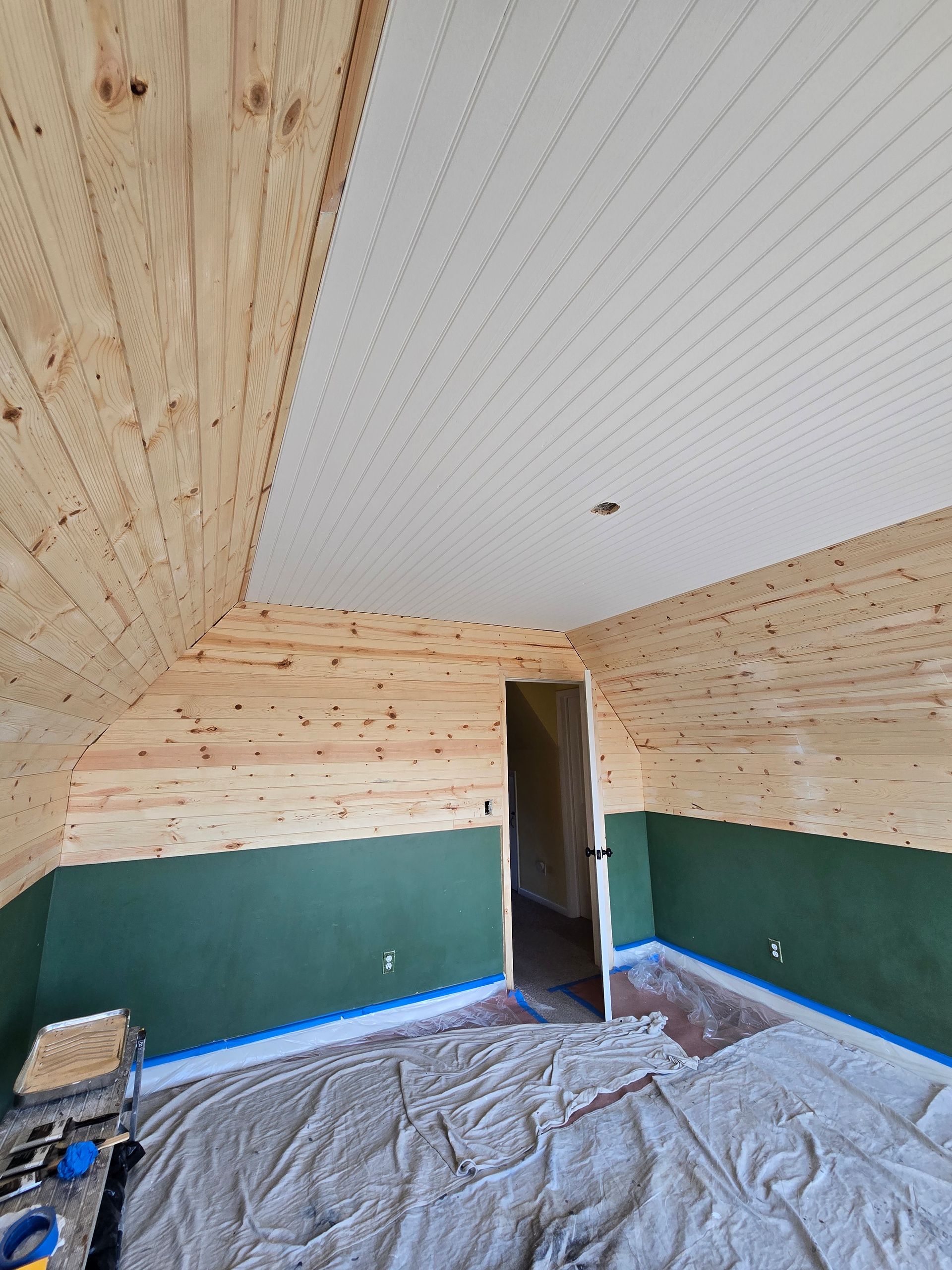 Empty attic room with sloped wood-paneled walls, white ceiling, open door, and covered floor.