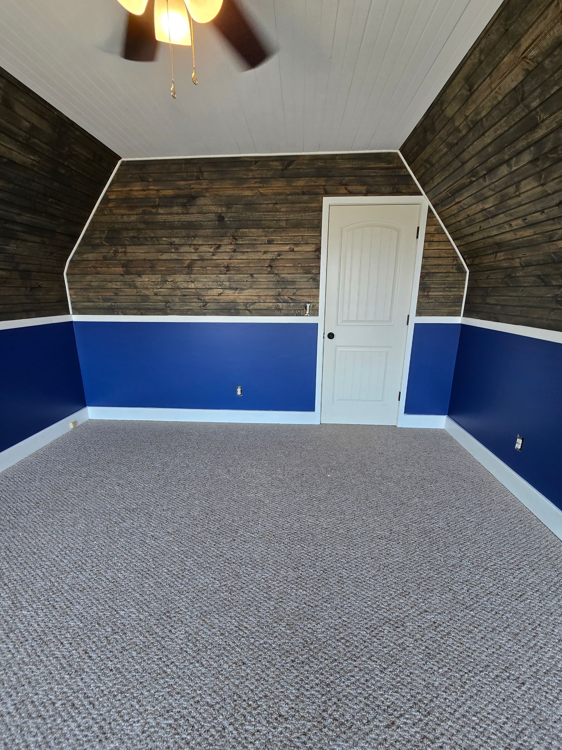 Empty room with gray carpet, blue lower walls, wood-paneled upper walls, and a white door.