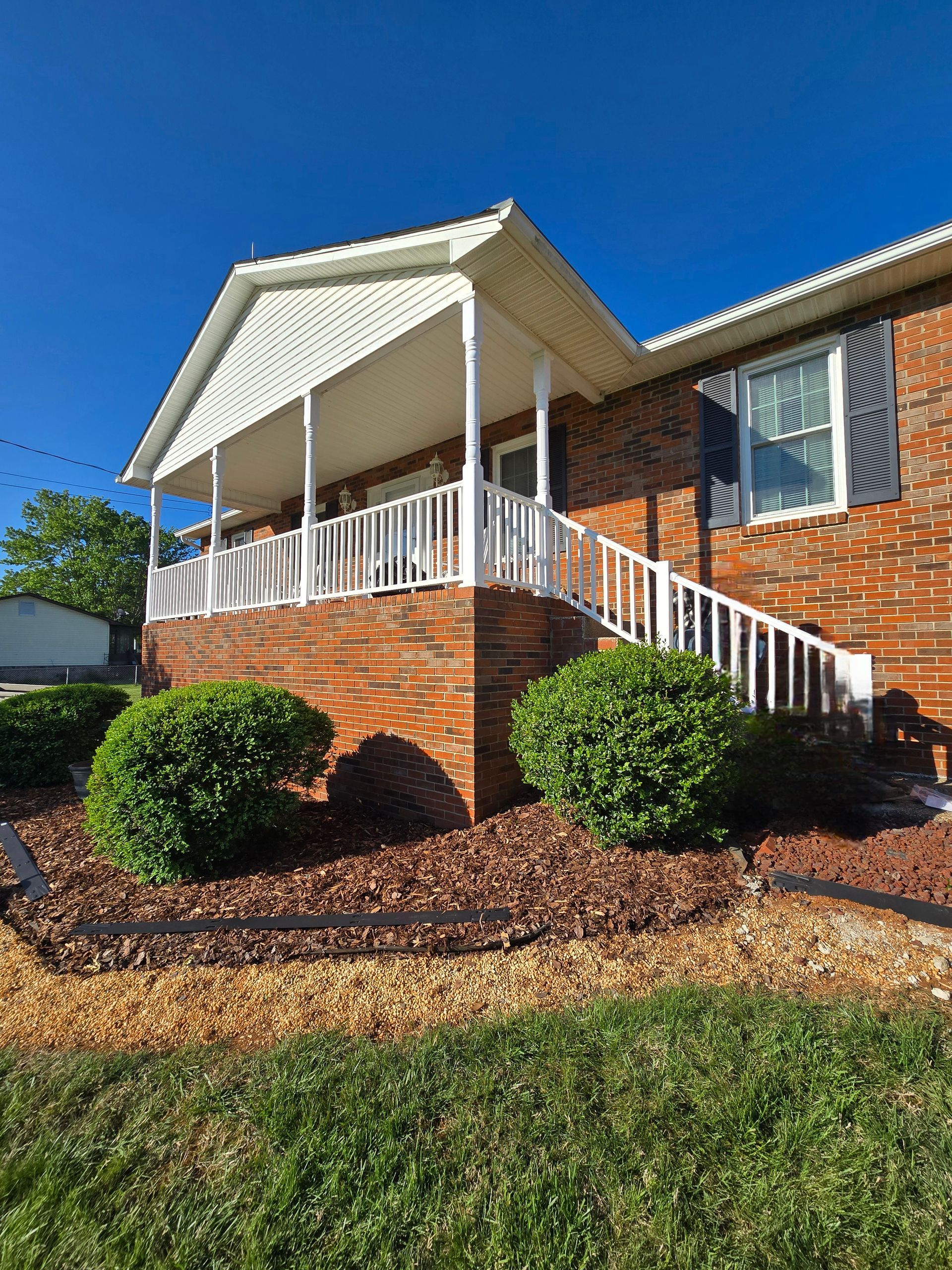 Brick house with white porch and stairs, landscaped shrubs, and a blue sky.