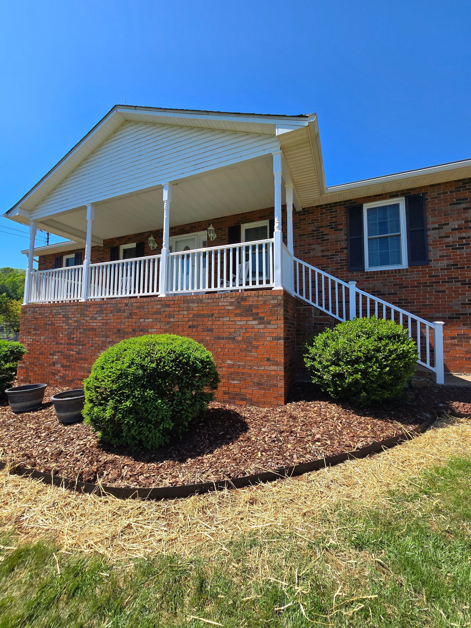 Brick house with white porch railings, steps, and trimmed shrubs under a bright blue sky