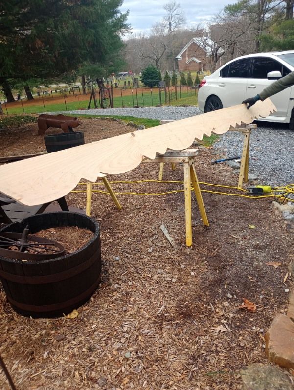 Long tan tarp stretched over yellow sawhorses in a yard beside a white car and barrel planter.