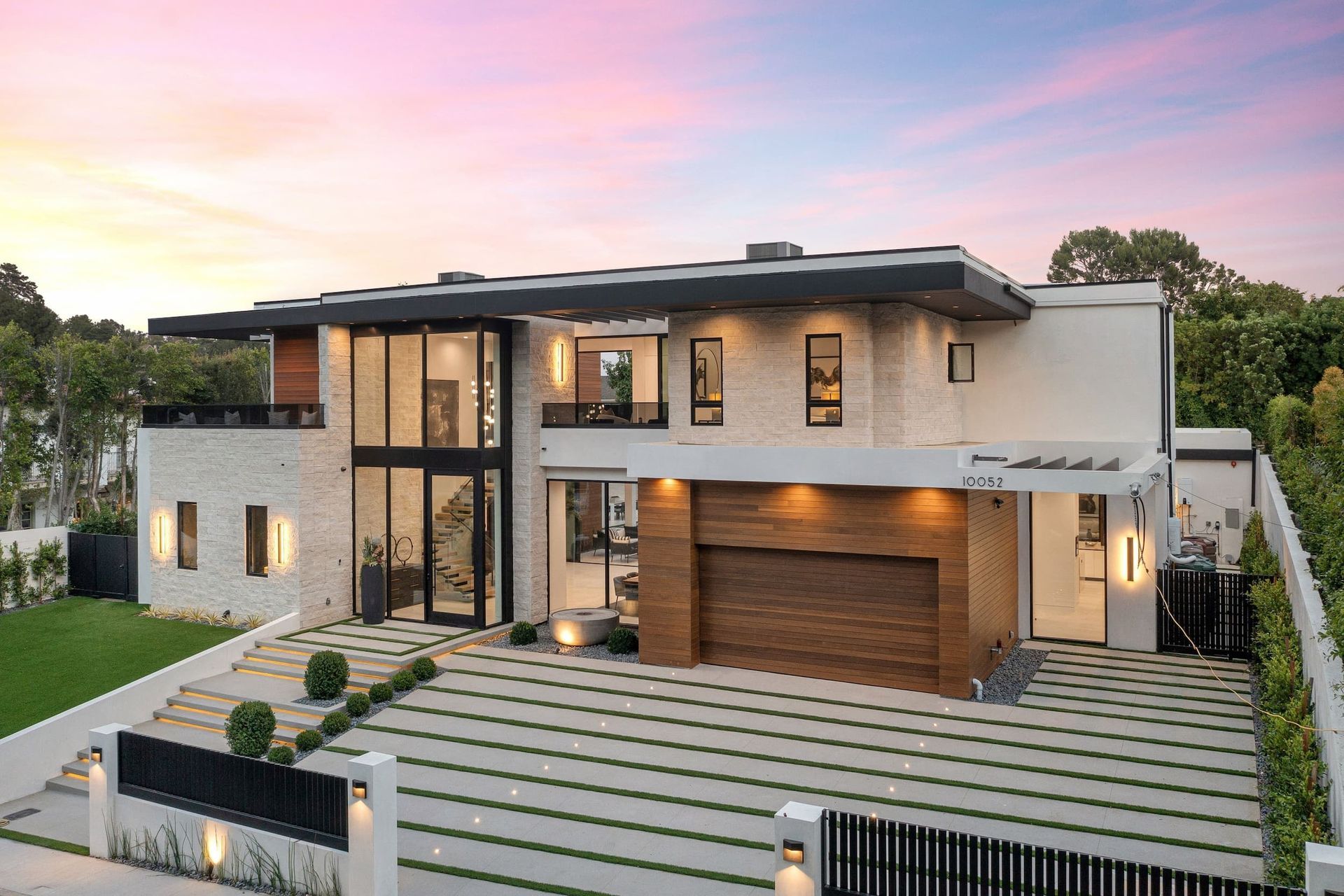 Modern two-story house with stone facade, glass windows, wooden garage door, and a paved driveway under a colorful sunset sky.