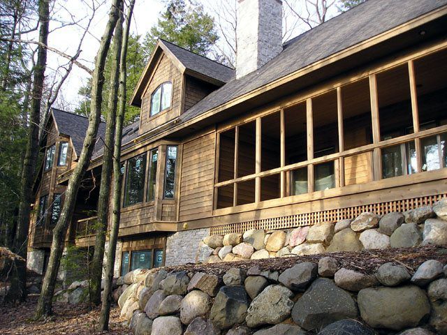 Wooden house with stone wall base, screened porch, windows, and chimney in a wooded setting.