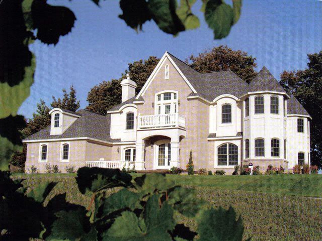 Two-story beige and white house with a gray roof, set on a green hill with a blue sky.