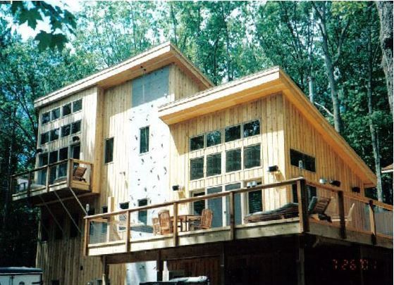 Two-story wooden house with large windows and a wraparound deck surrounded by trees.