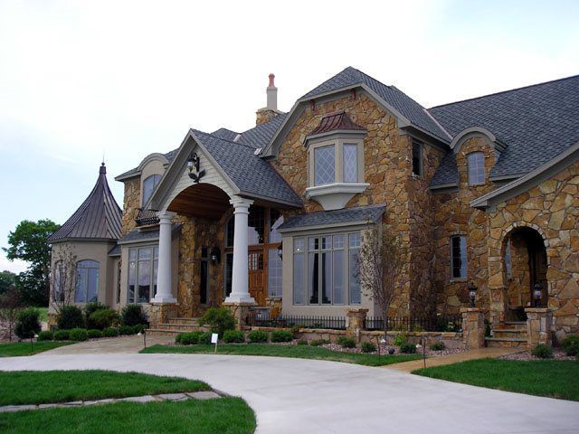 Stone-clad luxury house with columns, a curved driveway, and manicured landscaping under a cloudy sky.