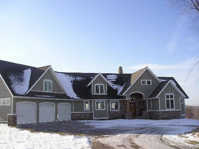 A large, gray house with snow-covered roof, three-car garage, and stone accents, under a blue sky.