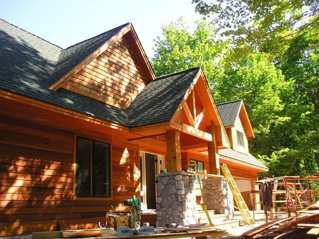 Wooden house under construction with stone pillar, cedar shake gables, and green surroundings.
