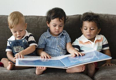 Three children sitting on a gray couch, each reading an open book with focused expressions.