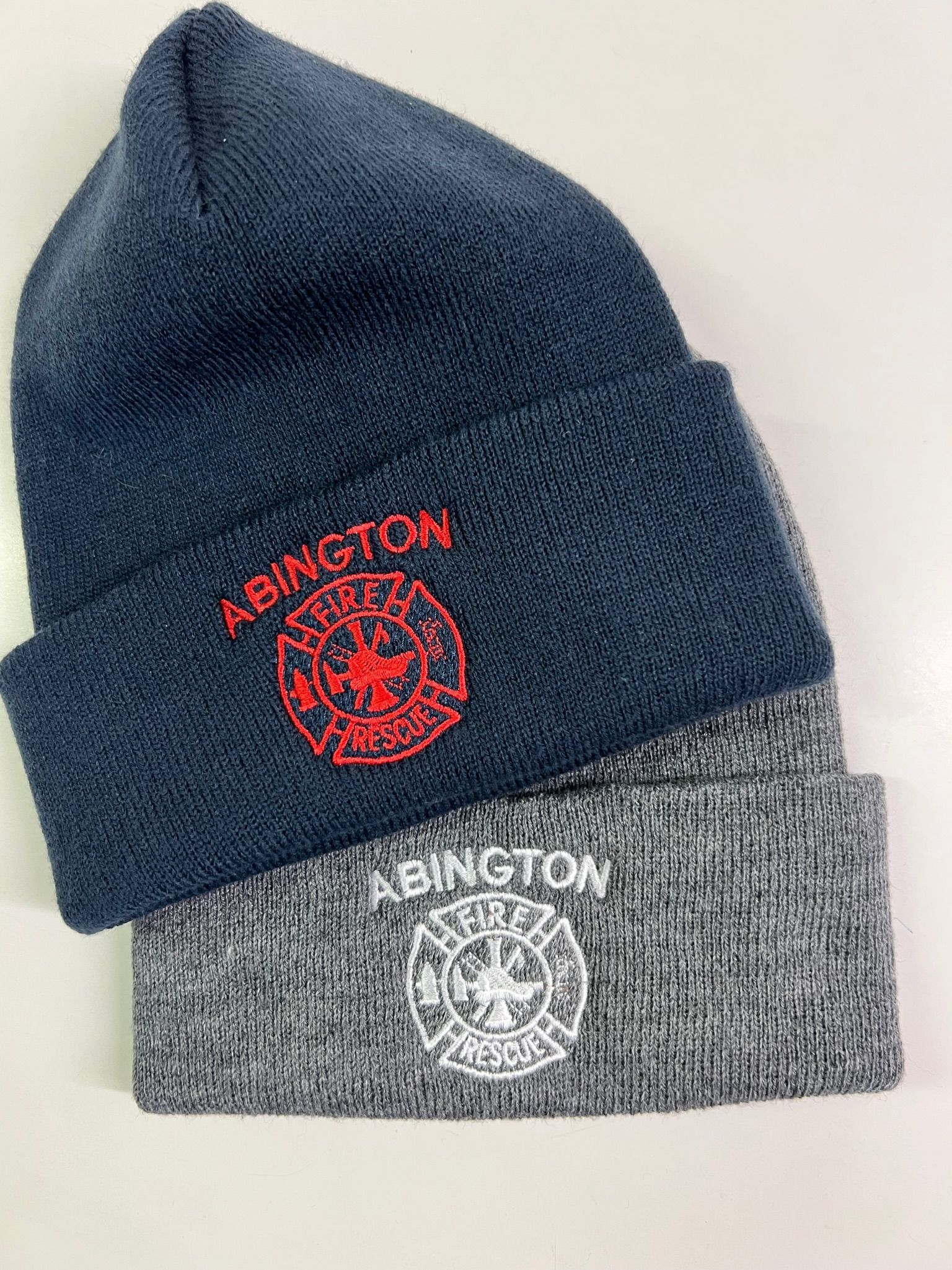 Two knit beanies, one navy blue with red embroidery and one grey with white embroidery, both featuring a fire emblem.