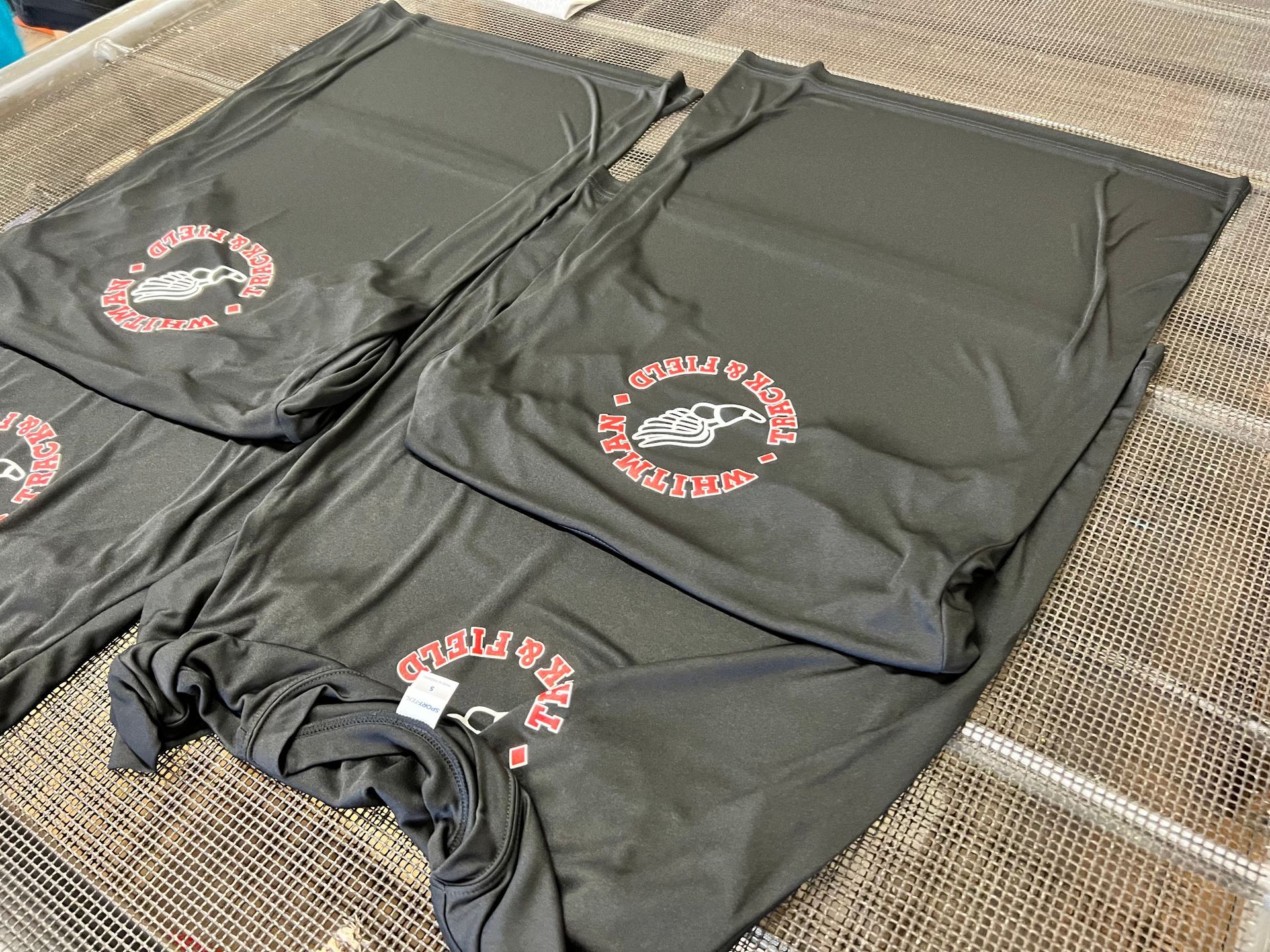 Black shirts with a custom silk screened red and white logo, laid on a textured surface.