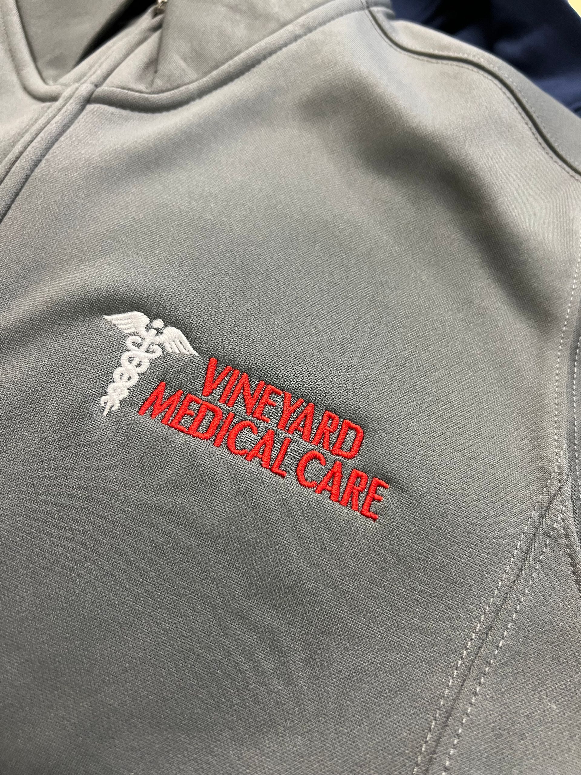 A grey fleece garment featuring an embroidered white caduceus symbol above the red text