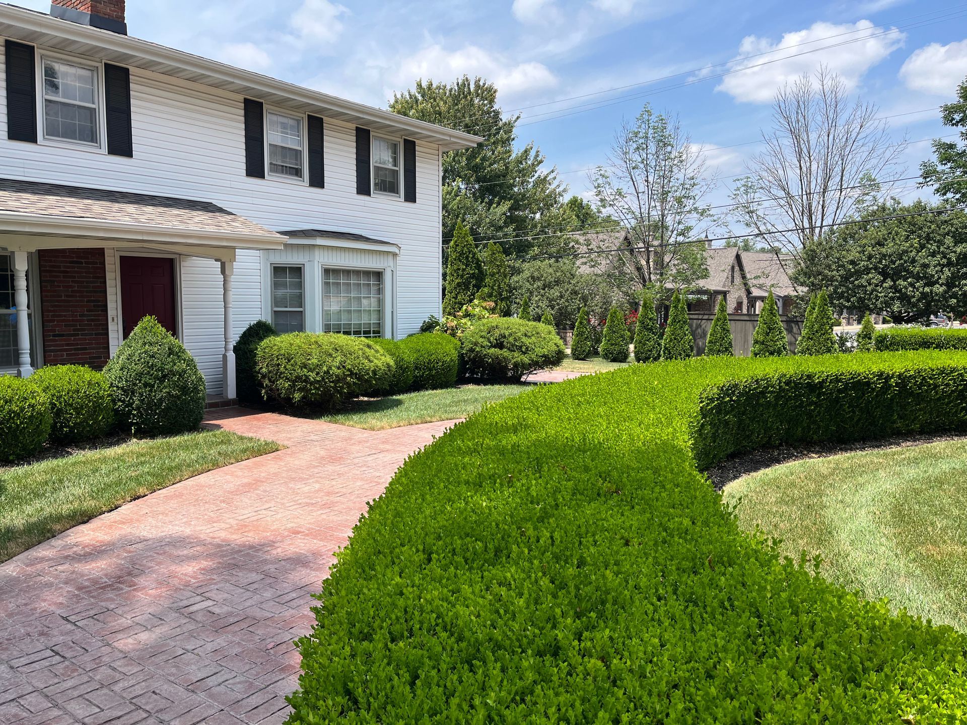 A white two-story house with a brick walkway and manicured green hedges on a sunny day.