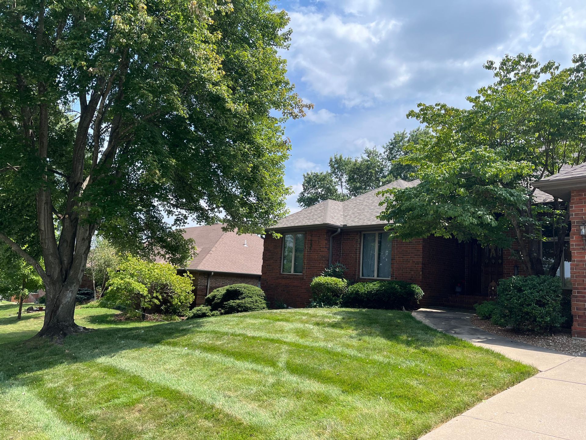 A single-story brick house with a green lawn and two large trees under a blue sky with light clouds.