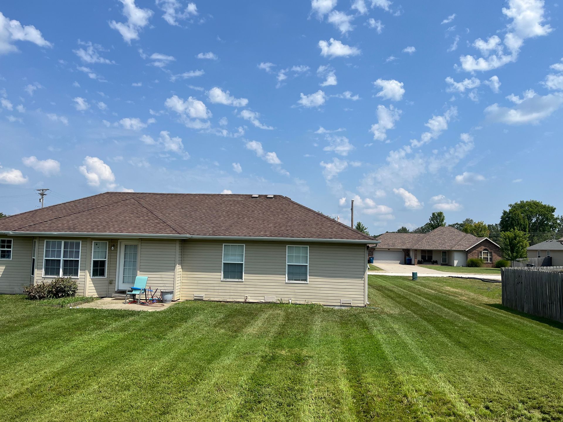 A tan, single-story house with a brown shingled roof and mowed lawn under a blue sky with scattered clouds.