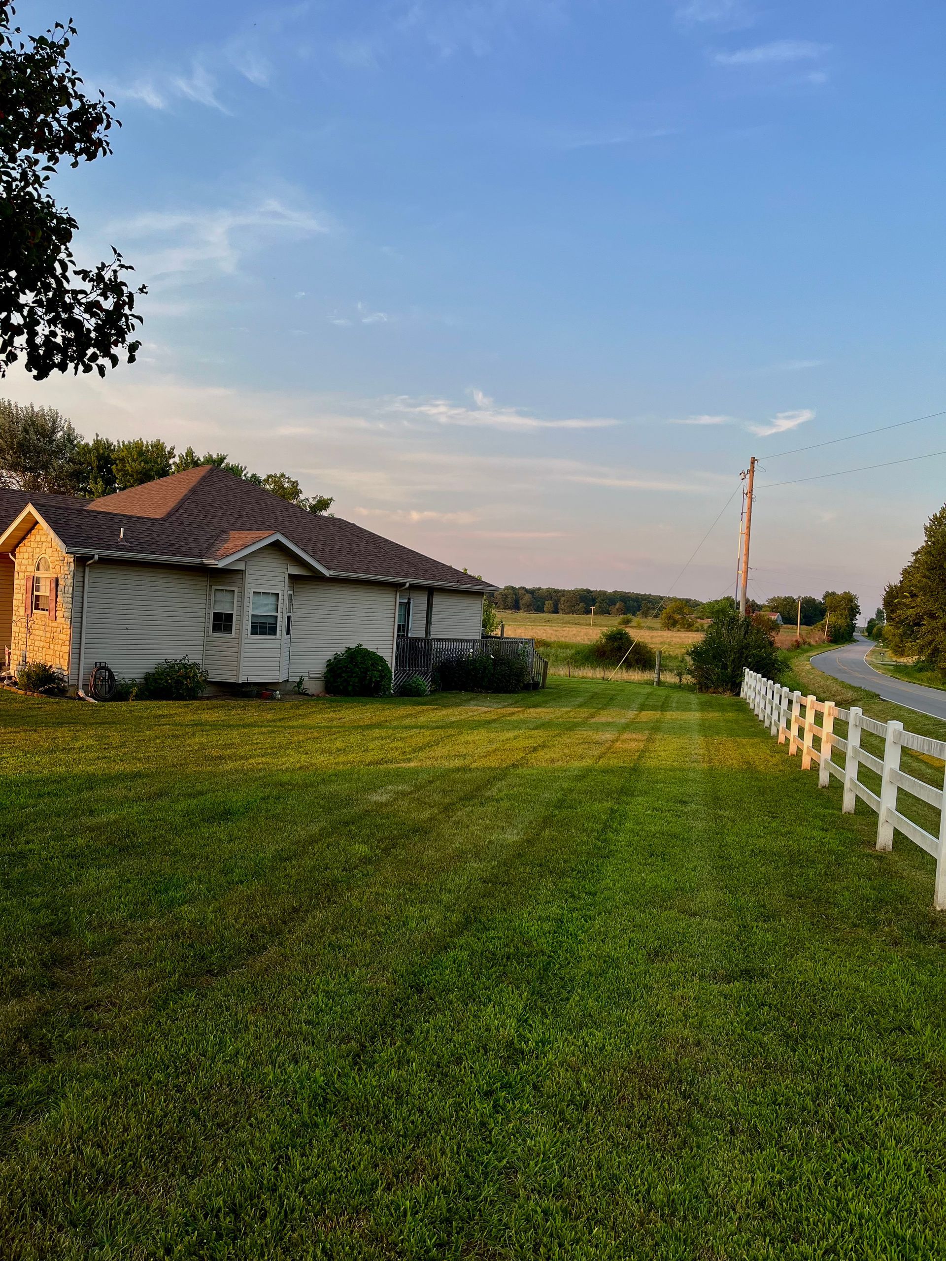 A light-colored house with a dark roof sits beside a white fence in a large, grassy field under a clear blue sky.