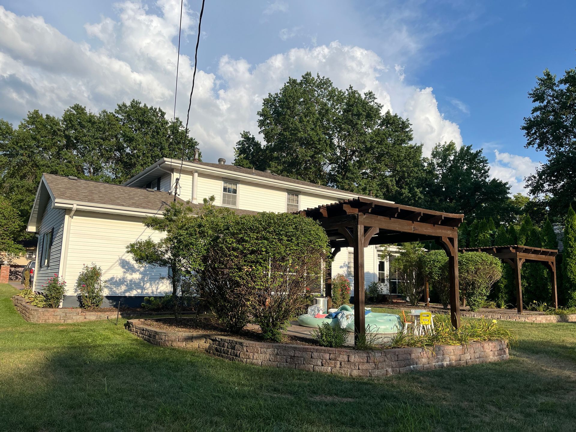 A light-colored house with a dark wooden pergola over a patio, surrounded by trees and a stone-walled garden.