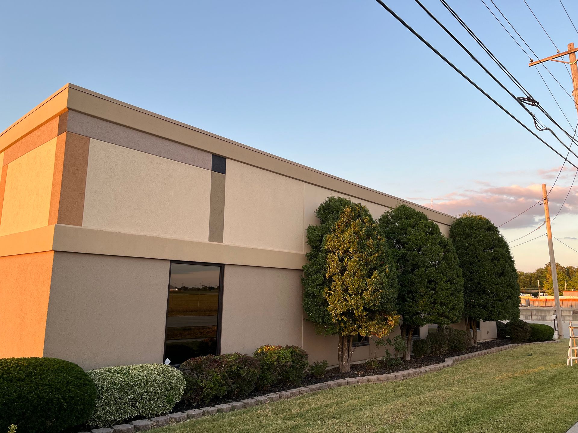 A long, low-profile beige commercial building with horizontal trim, a glass doorway, and manicured shrubs in front.