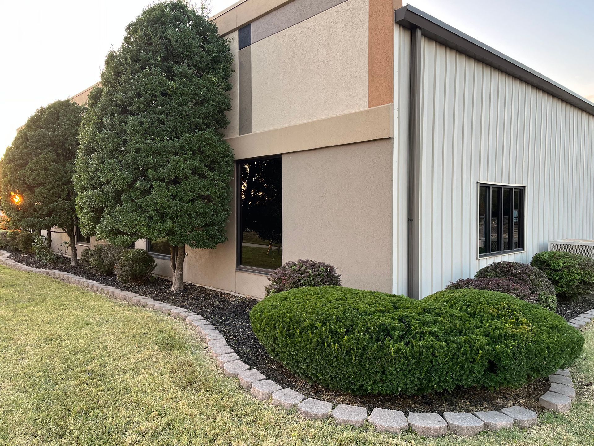 A tan commercial building with a metal side wall, landscaped with manicured shrubs and bushes along a curved stone border.