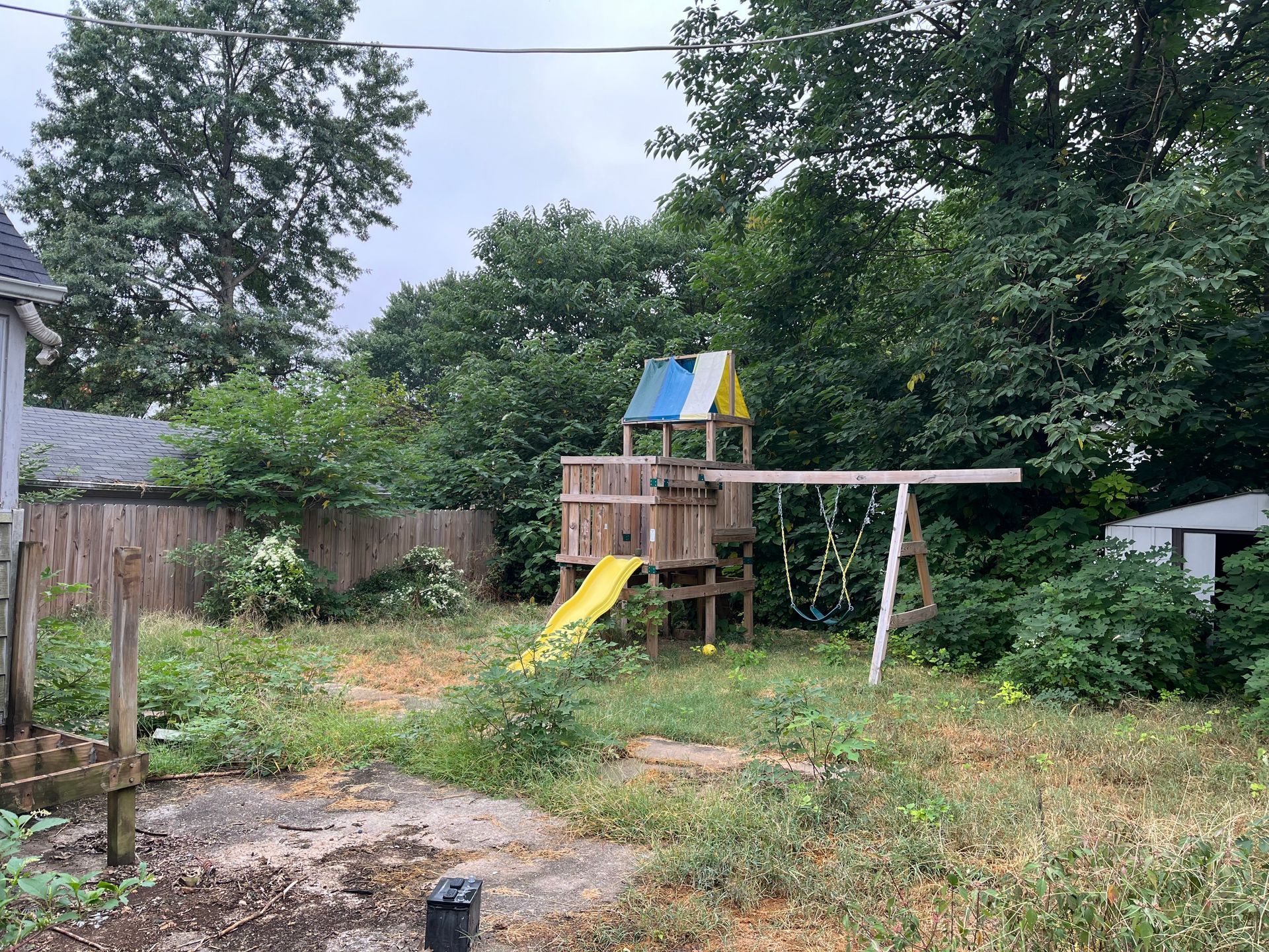 A wooden playset with a yellow slide and blue and yellow roof sits in an overgrown backyard with trees and a fence.
