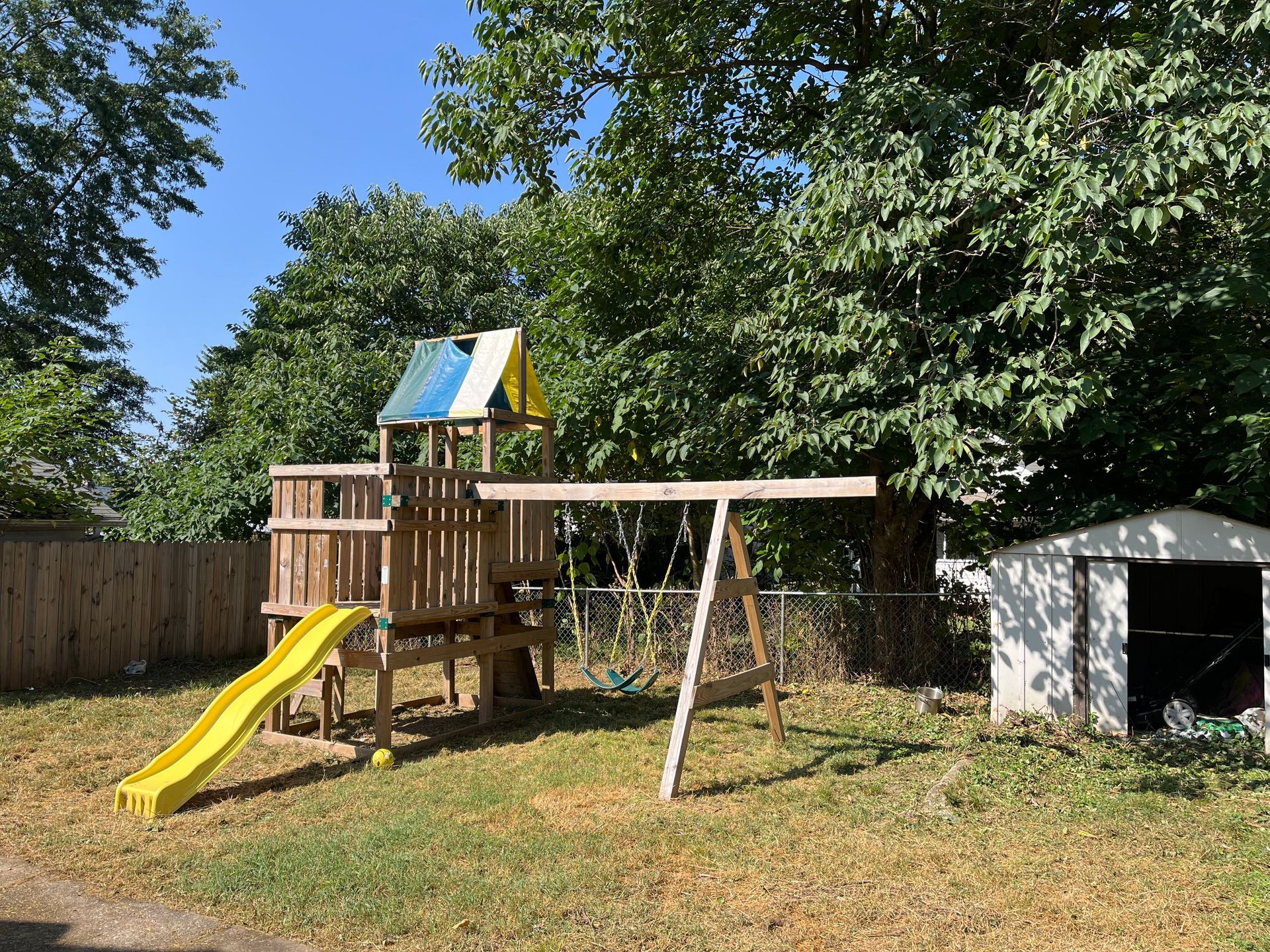 A wooden backyard playset with a yellow slide and swings, set in a grassy area next to a white shed and trees.