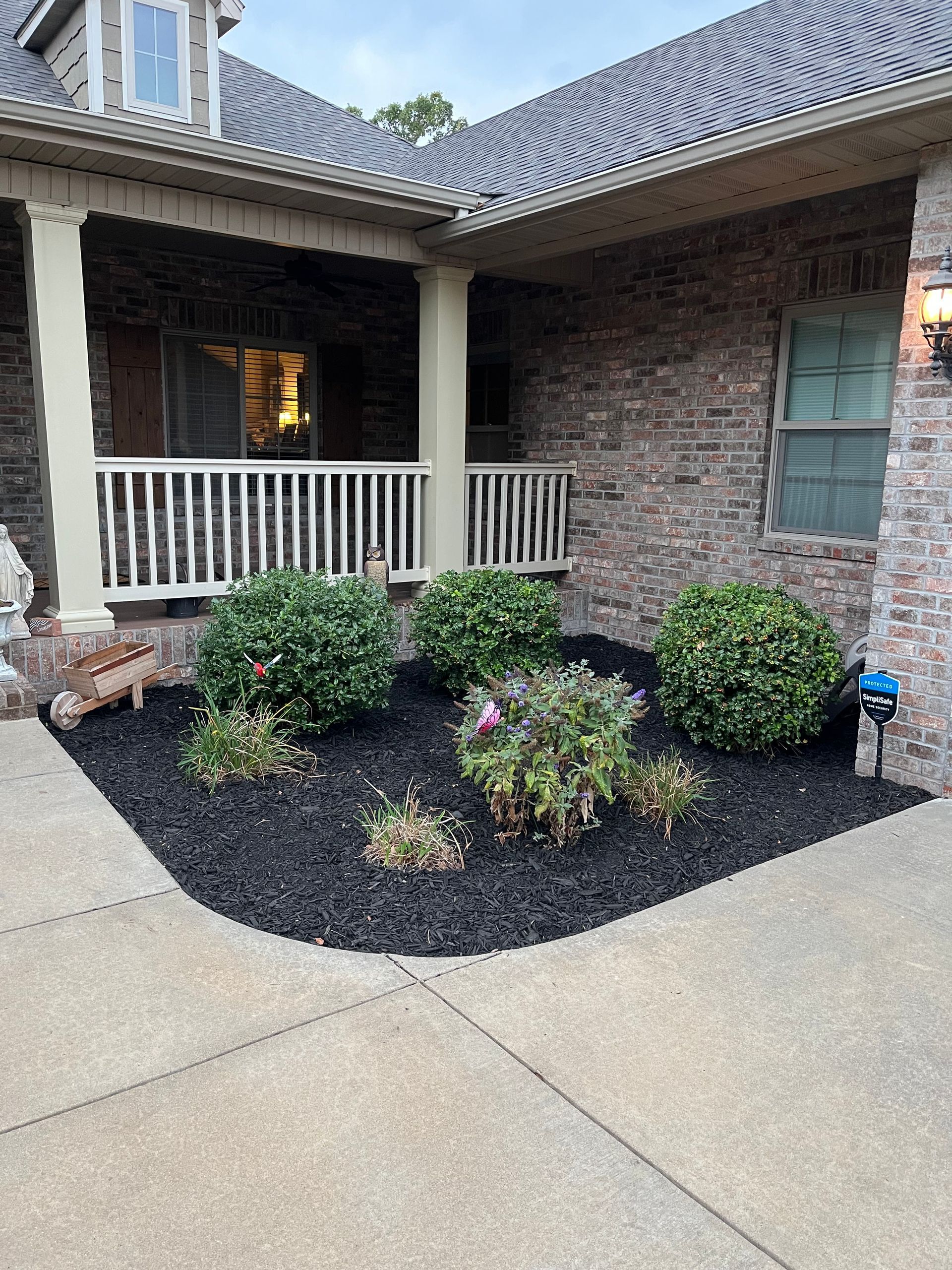 A brick house with a covered porch features a landscaping bed filled with black mulch, three round shrubs, and flowers.