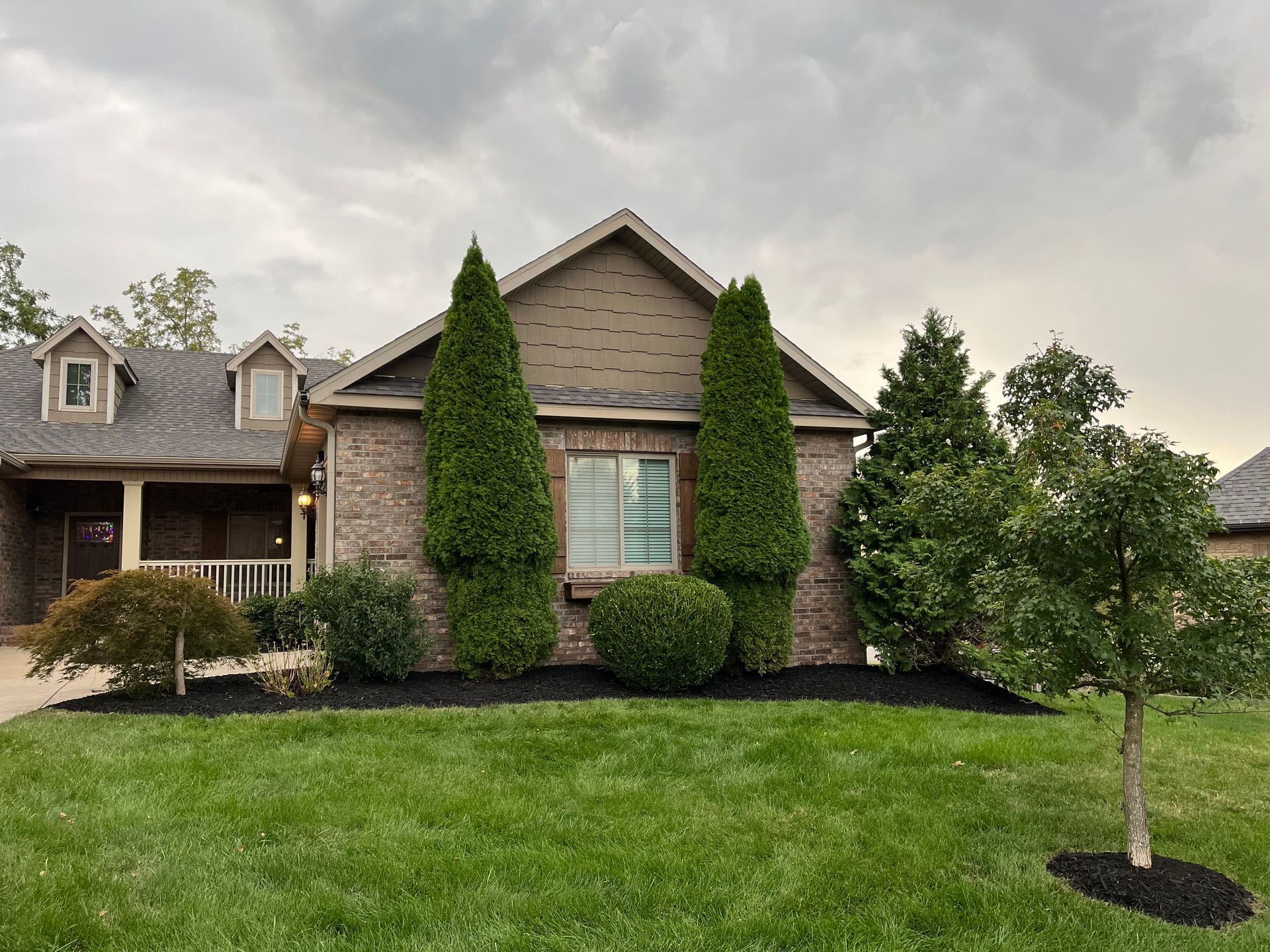 A front view of a brick house with a front porch, neat landscaping with tall evergreens, and a lawn under a cloudy sky.