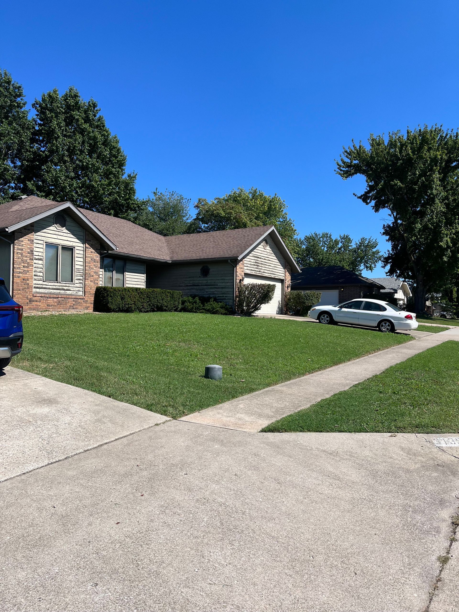 A brick house with a brown roof on a sunny day, with a white car parked on the street and a sidewalk in the foreground.