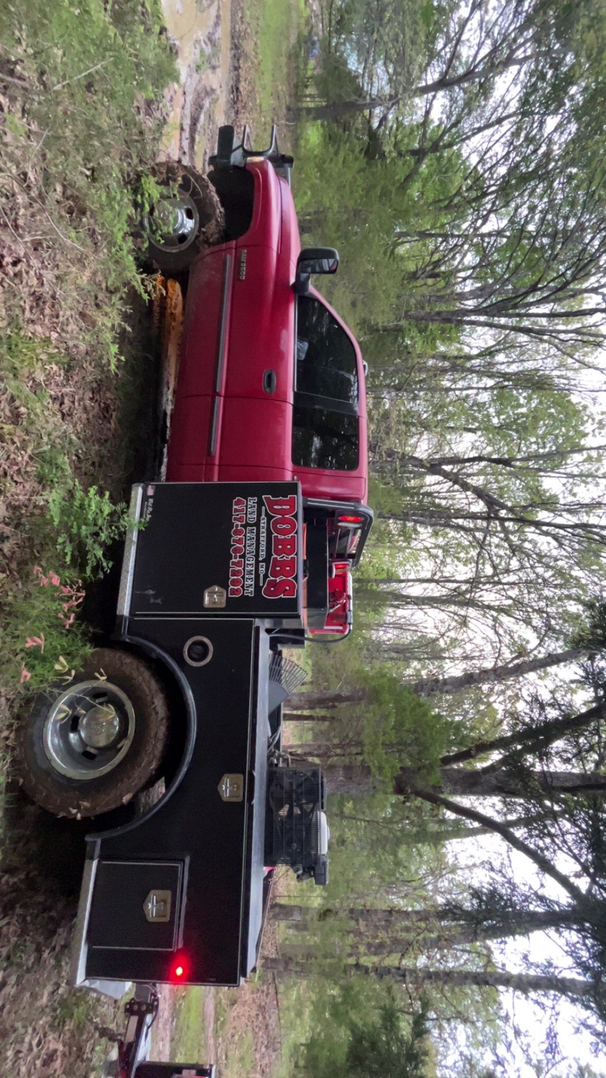 Red and black tow truck parked on a wooded roadside, hauling a red pickup.