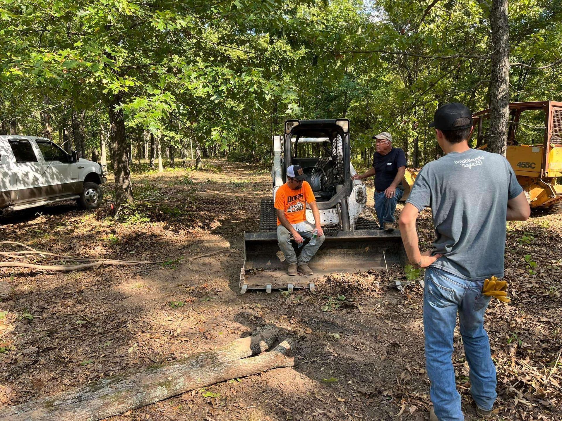 Two people talk in a wooded area with a white pickup truck, a skid steer, and yellow machinery nearby.