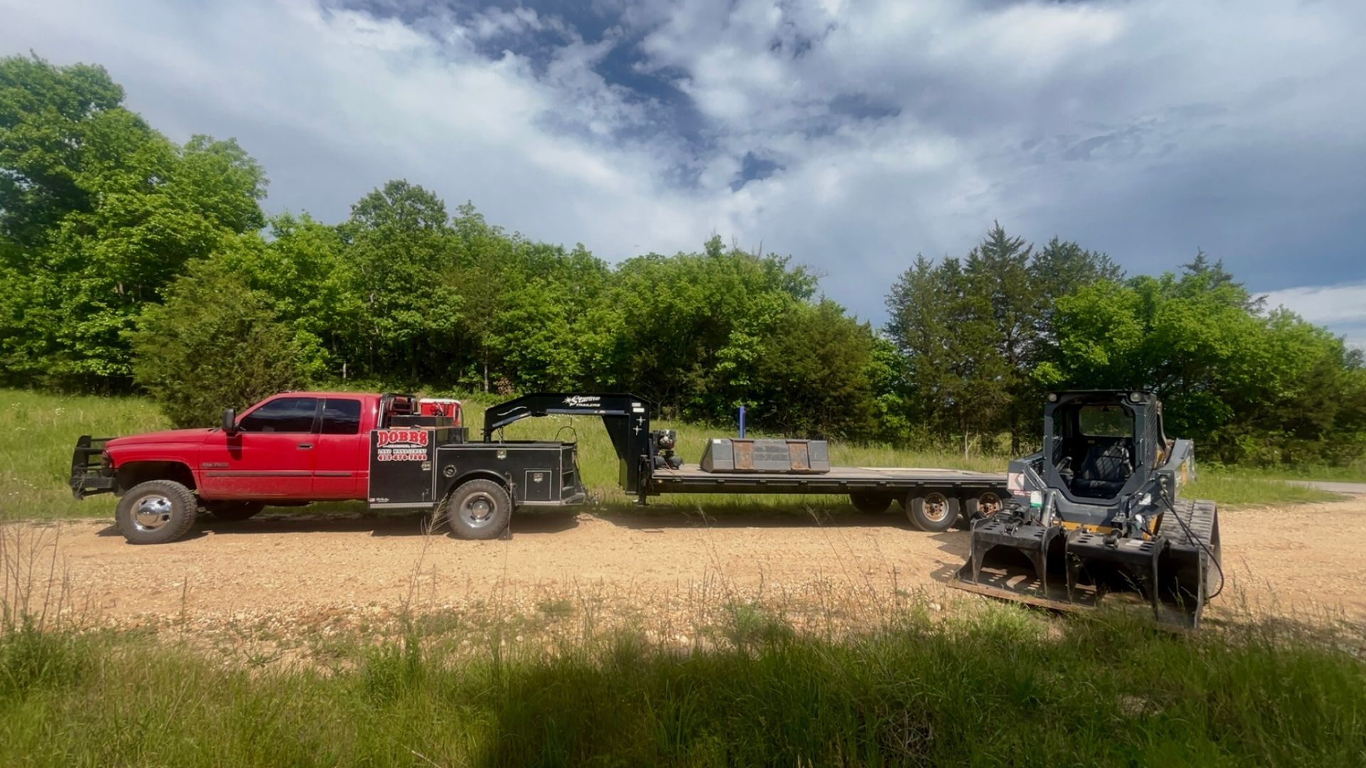 Red pickup truck towing a flatbed trailer with a skid steer in a grassy field under cloudy skies