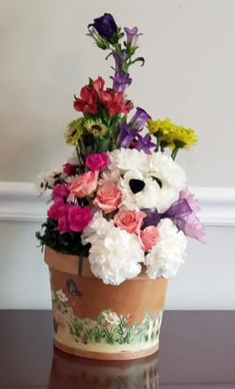 White teddy bear in a terracotta pot surrounded by colorful flowers against a plain wall