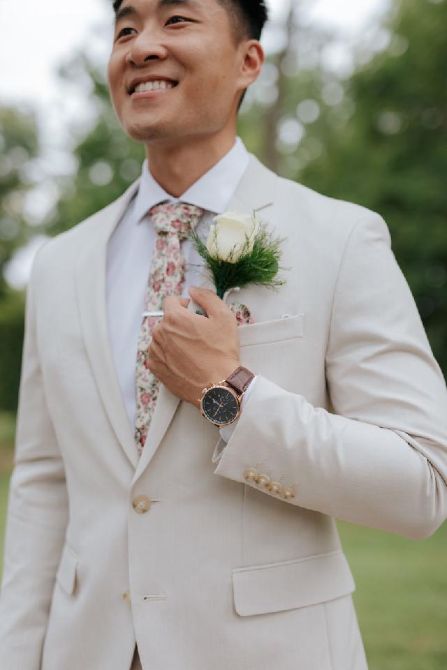 Man in a light suit smiling outdoors, adjusting his floral tie and boutonniere.