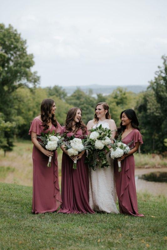 Four women in burgundy and white dresses holding flower bouquets outdoors on a grassy field.