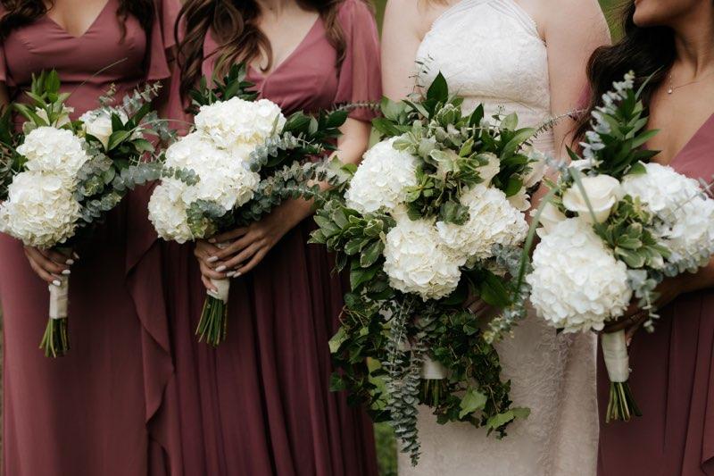 Bridesmaids in burgundy and ivory dresses holding white floral bouquets with greenery