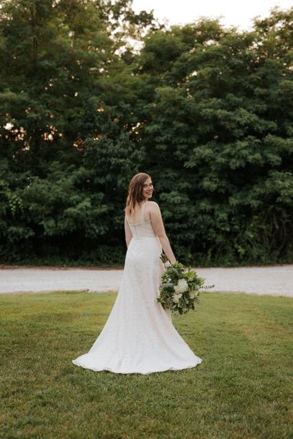 Bride in a white gown standing on a grassy lawn with a bouquet, backed by trees.