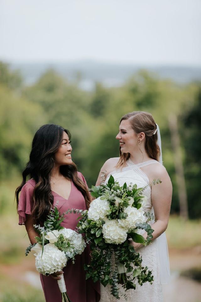 Two women in formal dresses smiling and holding bouquets outdoors on a blurred garden path