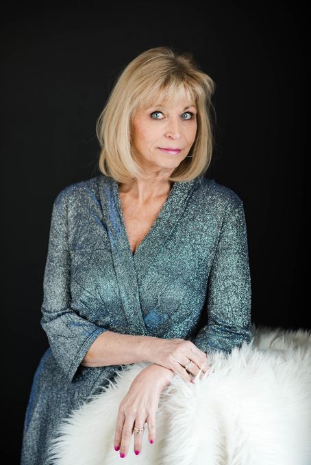 Woman in a teal patterned blouse seated against a dark background, with one hand resting on a white fur throw.