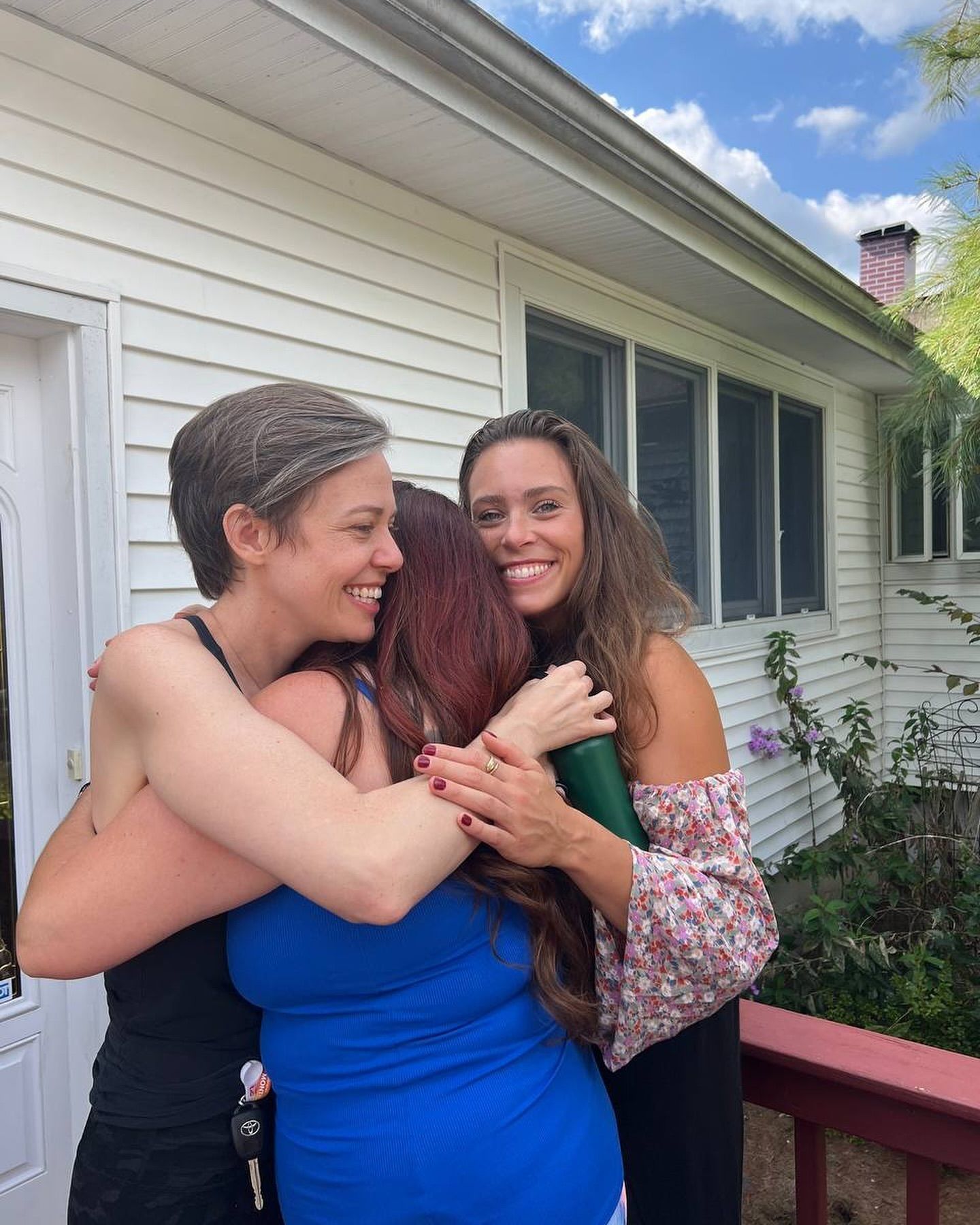 Three women are hugging each other in front of a white house.
