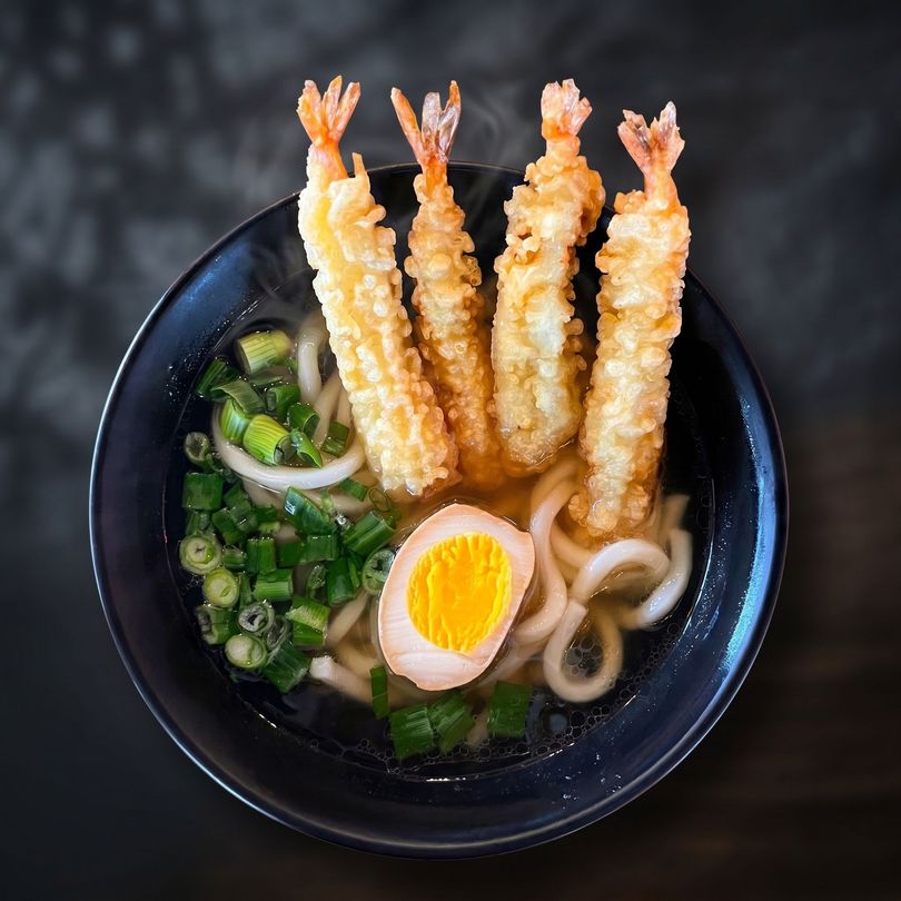 Bowl of ramen topped with three shrimp tempura, half a soft-boiled egg, and green onions
