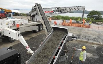 A man in a hard hat is standing in front of a conveyor belt filled with concrete.
