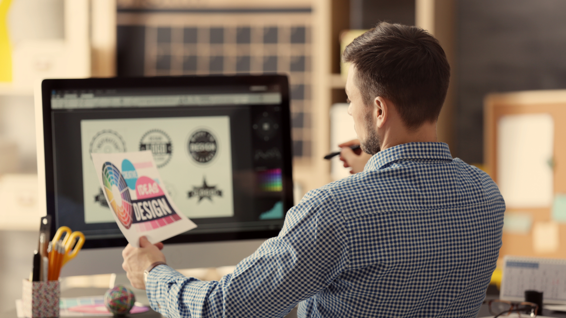 Graphic designer at desk compares print design to digital version on computer screen.