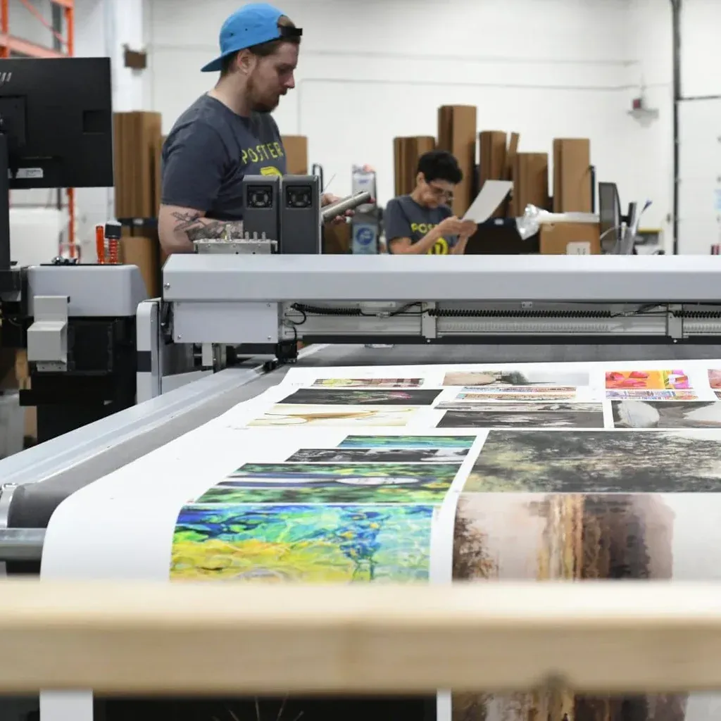 People operating a large format printer. A worker in a blue cap watches as the machine prints. A young person looks at papers.