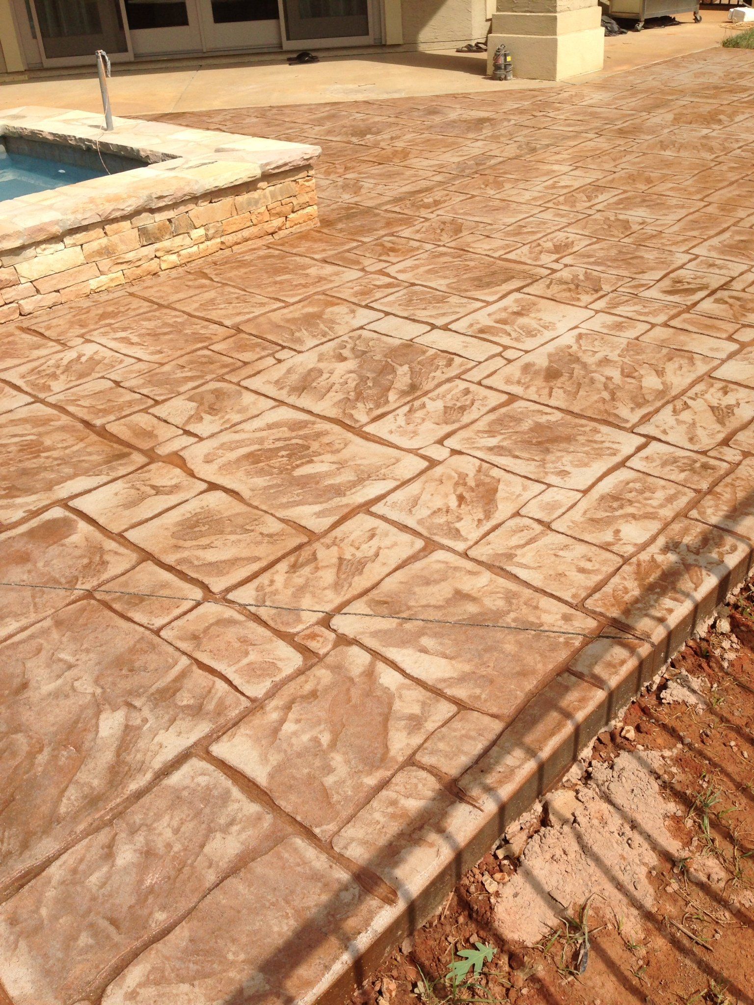 Stamped concrete patio beside a pool, with tan textured paving and brick edging.