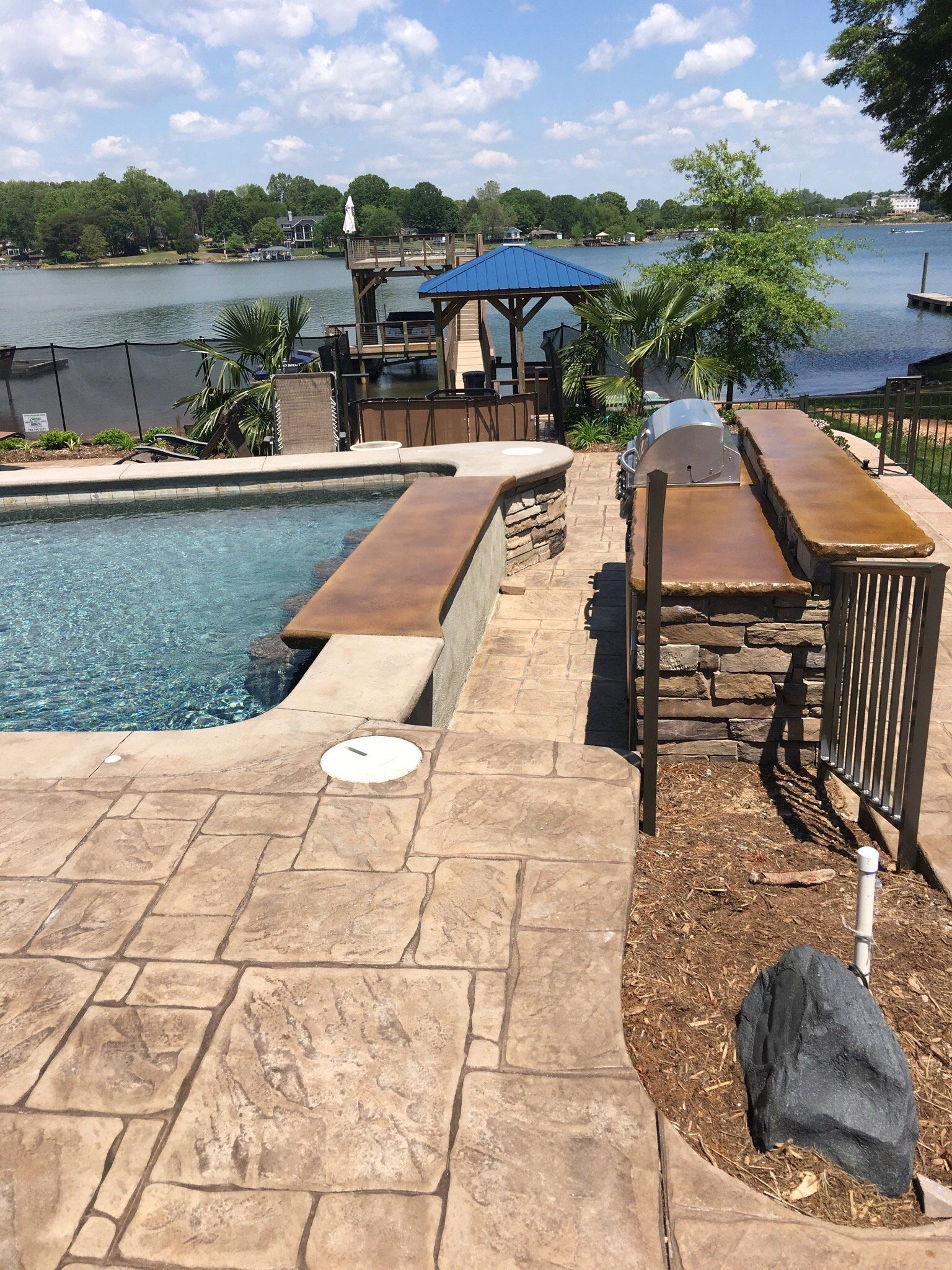 Poolside patio beside a lake, with lounge chairs, umbrellas, and a stone walkway under a blue sky.