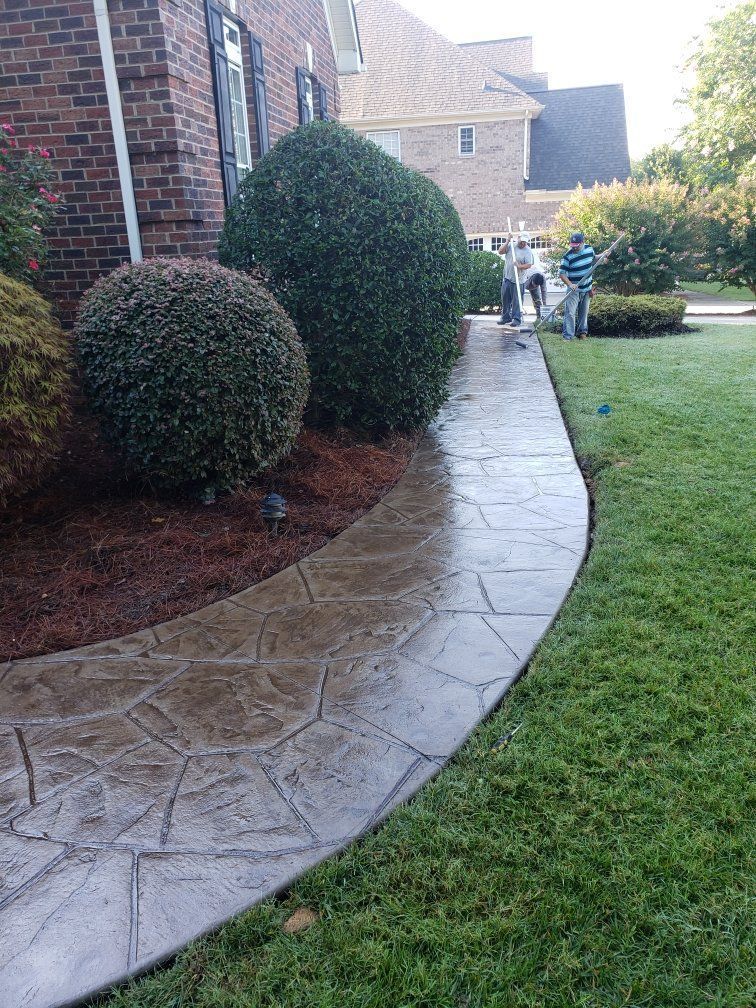 Curved stone walkway beside brick houses, trimmed shrubs, and a green lawn in a residential yard
