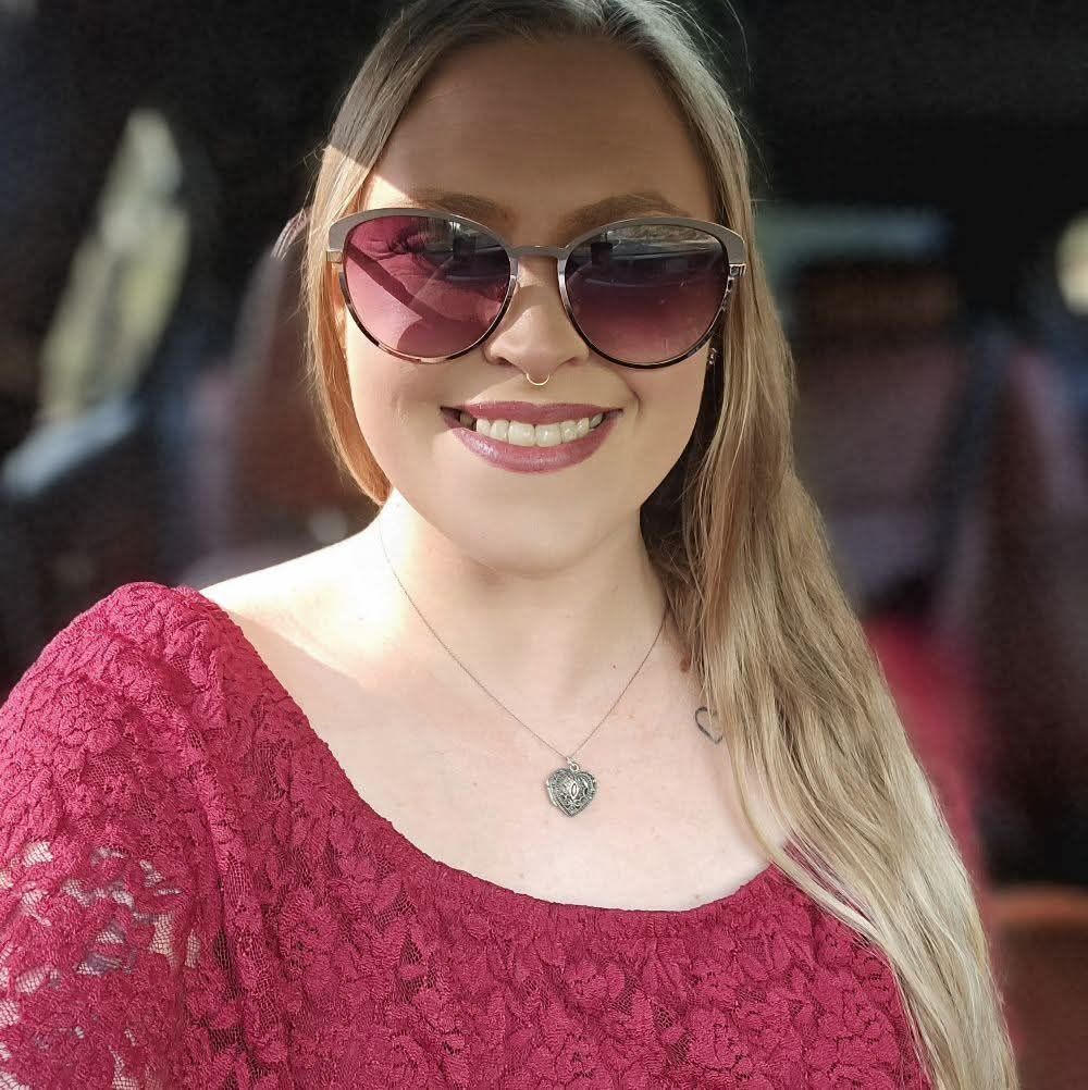 Woman with long blonde hair, wearing sunglasses, a red lace top, and a heart pendant, smiling.