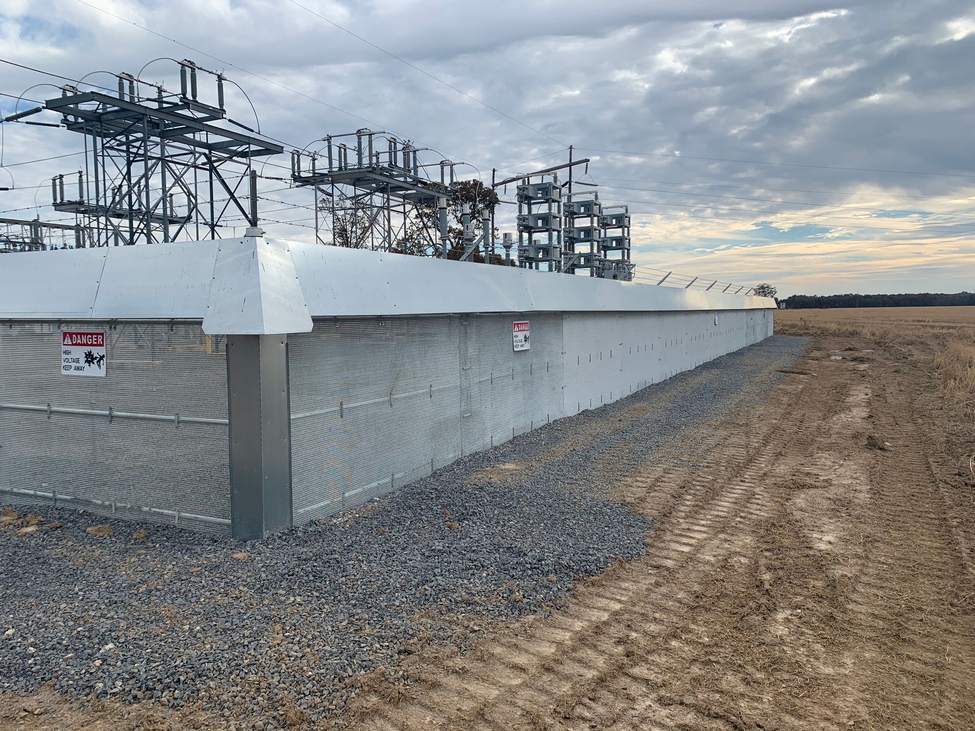 Electrical substation equipment behind a white barrier beside a gravel road under a cloudy sky