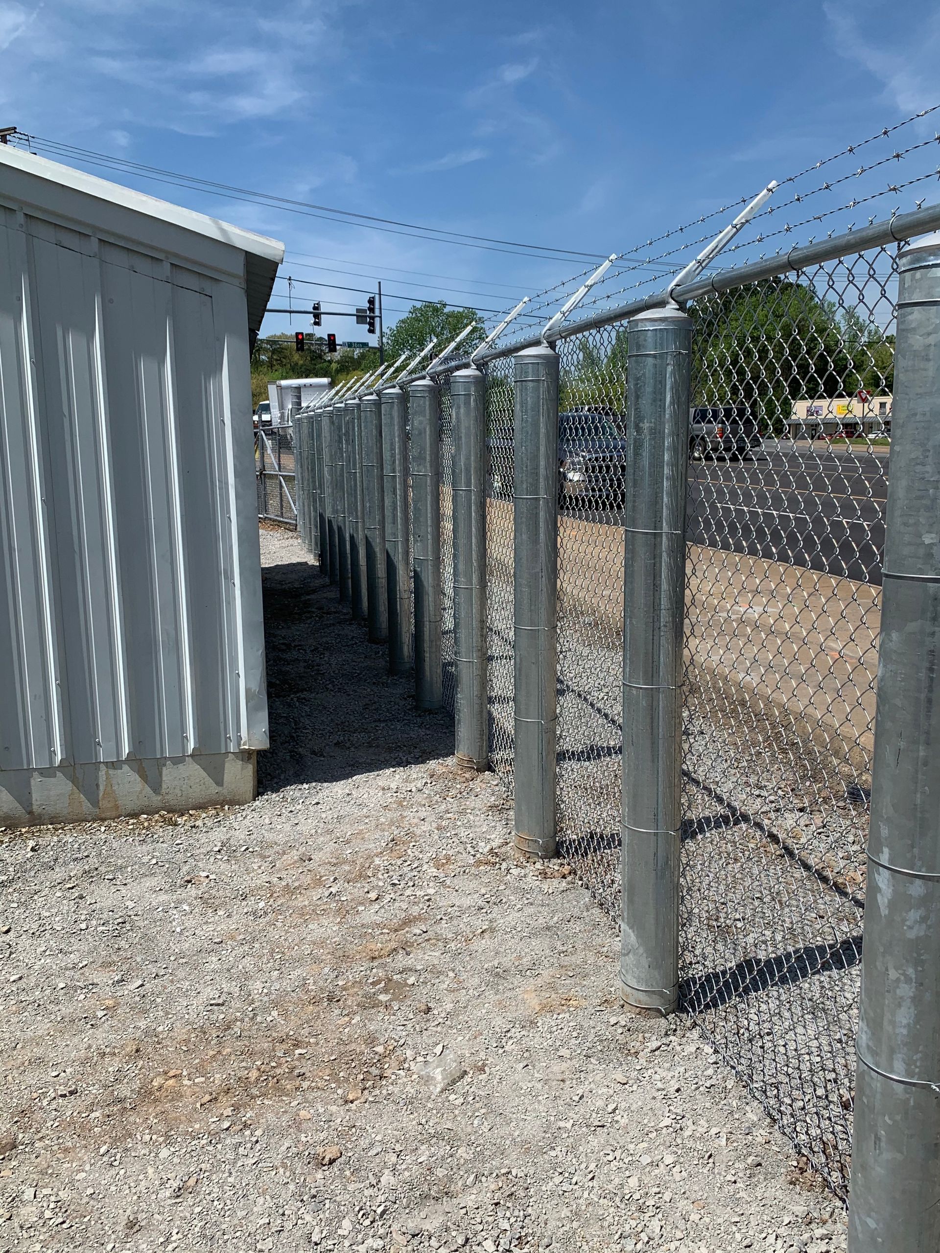 Chain-link fence with barbed wire beside a corrugated metal shed on a gravel path under a blue sky